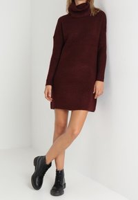 Vestito in maglia color marrone scuro con colletto a scaldacollo, maniche lunghe, spacchi laterali e tessuto testurizzato. Abbinato a bottini neri con lacci.