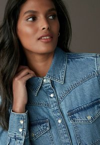 Veste en denim bleu clair avec un col pointu, deux poches avant, des boutons métalliques et un tissu texturé. Modèle tenant le col d'une main.