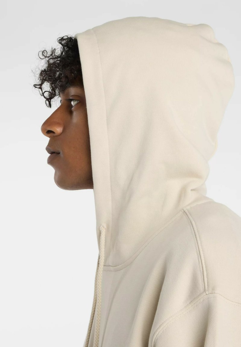 Beiger Hoodie mit Kordelzug und großer Kapuze. Weicher Stoff und sichtbare Nähte entlang der Nähte. Seitenansicht.