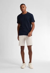 T-shirt bleu marine à manches courtes, col rond, porté avec un short beige clair et des baskets blanches. Design simple avec des détails subtils sur l'ourlet du t-shirt.