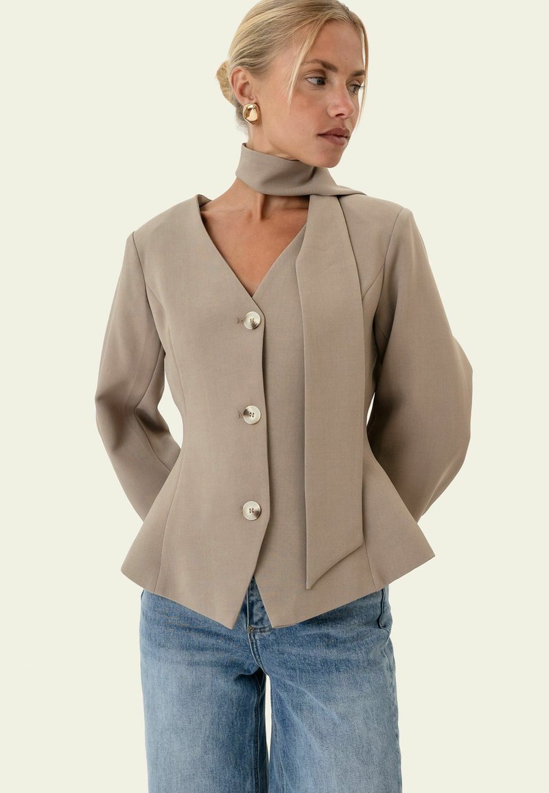 Femme portant une veste beige cintrée avec de gros boutons et un détail d’écharpe attaché, associée à un jean bleu, regardant sur le côté.
