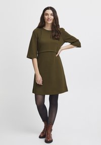 Olivgrön klänning med texturerad yta, korta ärmar och figurnära liv. Buren med svarta strumpbyxor och bruna ankelstövlar.