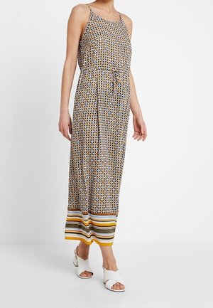 Kobieta ubrana w bezrękawnikową, długą sukienkę z geometrycznym wzorem i paskiem w paski na dole, w połączeniu z białymi, otwartymi sandałami na obcasie.