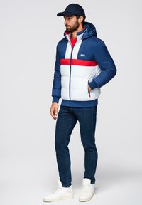 Padded hooded jacket w kolorze granatowym i białym, z czerwonym paskiem. Noszona na czerwonej koszuli, w zestawieniu z ciemnymi dżinsami i białymi trampkami.