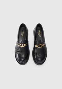 Black leather loafers with a fringe design and gold chain accent. Smooth texture with a round toe and thick black sole.