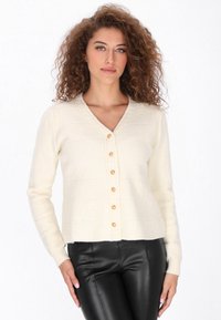 Cremfarbener Strickcardigan mit V-Ausschnitt, strukturierten horizontalen Streifen und sechs goldenen Knöpfen auf der Vorderseite. Über schwarzen Lederhosen getragen.