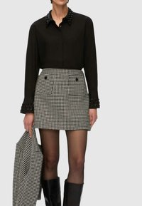 Camicia nera con bottoni e colletto decorato con perline, abbinata a una gonna pied de poule bianca e nera con tasche frontali e vestibilità aderente. Stivali neri al ginocchio.