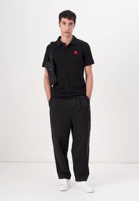 Μαύρο polo μπλουζάκι με διακριτό κόκκινο λογότυπο, σε συνδυασμό με φαρδιά μαύρα παντελόνια και λευκά αθλητικά παπούτσια. Περιλαμβάνει μια μικρή μαύρη χιαστί τσάντα.