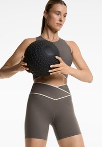Sport bra s visokim vratom i hlačice visokog struka u prigušenoj smeđoj boji s kontrastnim bež šavovima. Drži teksturiranu, crnu medicinsku loptu.