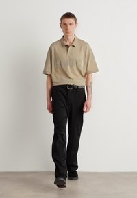 Beige polo-skjorte med preget "HUGO" logo, krage, korte ermer, kombinert med svarte rette bukser og belte, båret med svarte sko.