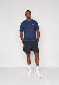 Blå Nike t-skjorte, svarte shorts, hvite sokker og sportsko. Korte ermer, enkelt design og logoakenter. Modell som står oppreist.