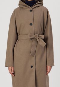 Long manteau à capuche en tissu marron doux. Comprend une ceinture nouée à la taille et de gros boutons noirs, avec des manches à épaule tombante.