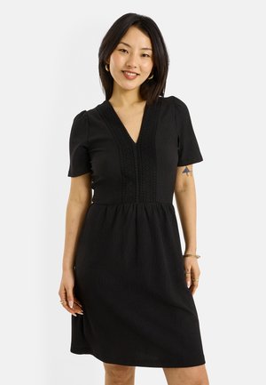 Mujer con cabello negro hasta los hombros, vistiendo un vestido negro de manga corta y cuello en V, sonriendo, con pendientes de aro dorados y anillos.