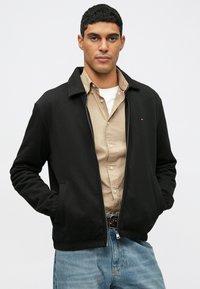 Crna jakna na zatvaranje sa ovratnikom i bočnim džepovima, s logotipom na prsima. Nosi se preko bež košulje na dugmad i traperica.