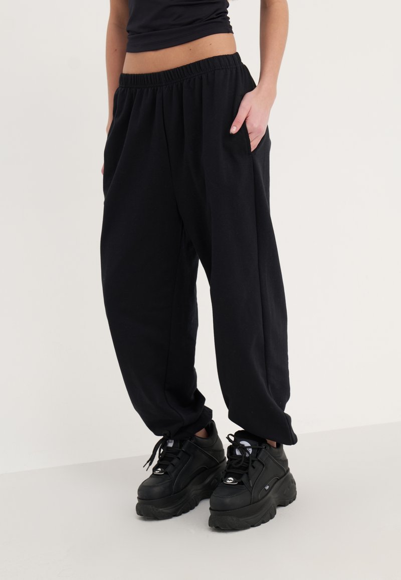 Pantalons de survêtement noirs avec une taille élastique, poches latérales et une coupe ample, associés à des baskets noires chunky. Texture lisse.