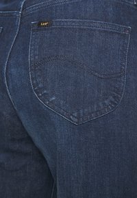 Detailní záběr na kapsu tmavě modrých džín s jemným vlnkováním a malou černou etiketou značky Lee.