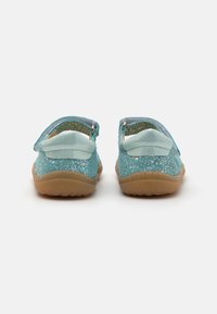 Sandalias brillantes en tono azul verdoso con una suave correa azul, soporte trasero y una suela de goma flexible en color marrón claro. La vista trasera muestra una forma redondeada y una superficie texturizada.