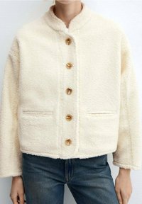 Veste en polaire de couleur crème avec col montant, six gros boutons marron et deux poches avant ; présente un tissu doux et texturé.