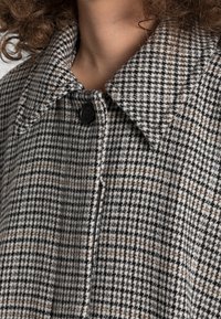 Manteau à motif pied-de-poule gris et noir avec un col pointu, doté d'un bouton noir et d'un tissu texturé.