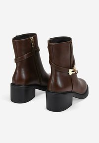 Botas de piel marrón hasta el tobillo con un tacón plano de goma negra, cremallera lateral y una correa decorativa con acentos metálicos. Textura suave, punta redondeada.