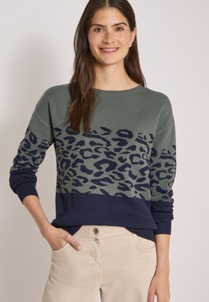 Kvinde med langt brunt hår iført en grøn og mørkeblå leopardmønstret sweater og beige bukser, smilende med den ene hånd i lommen.