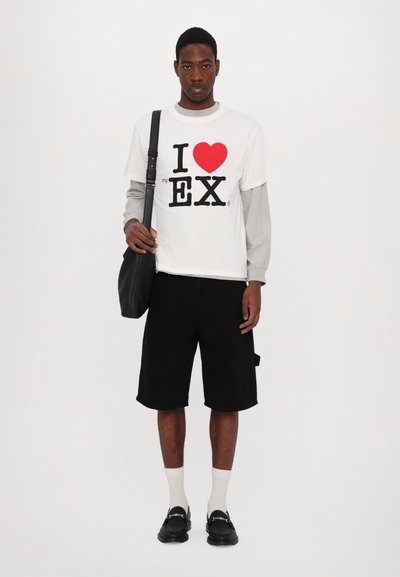Λευκό T-shirt με μαύρο και κόκκινο γραφικό κείμενο, γκρι μακρυμάνικο στρώμα από κάτω, μαύρες βερμούδες, μαύρη τσάντα, λευκές προμήθειες και μαύρα παπούτσια.