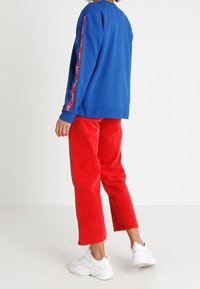 Blå sweatshirt med röd randdetalj längs ärmen, tillsammans med röda manchesterbyxor och vita sneakers.