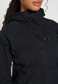 Czarna kurtka typu softshell z wysokim kołnierzem, zapięciem na zamek z przodu oraz teksturowanym materiałem z przeszytymi akcentami i regulowanym kapturem.