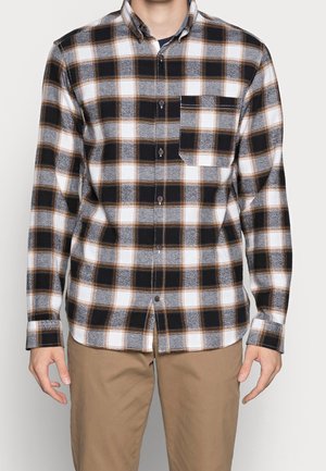 Man wearing long-sleeve buttoned black, white, gray, and brown plaid shirt with chest pocket and beige pants against a white background.