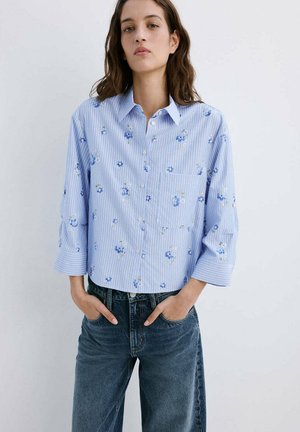 Femme portant une chemise boutonnée à manches longues bleu clair à motifs floraux et rayures, avec les poignets relevés et un jean bleu foncé, les mains dans les poches.