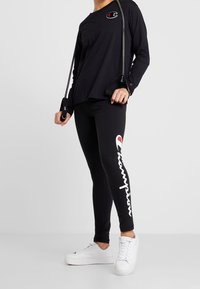 Maglietta nera a maniche lunghe con dettagli del logo, abbinata a leggings neri con branding bianco. Le scarpe sono bianche e viene tenuto uno strumento per il fitness.
