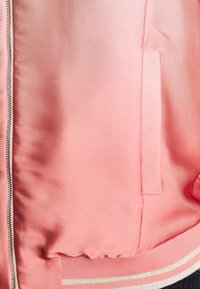 Roze satijnen bomberjack met een gladde textuur, voorzien van een zilveren ritssluiting, zijzak en geribbelde manchetten met twee gouden strepen.