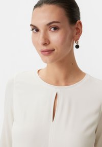Weißes Blusenshirt mit einem Schlüsselausschnitt und langen Ärmeln. Model trägt goldene und schwarze Hängehänger. Teint ist hell.