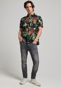 Maglietta nera a maniche corte con un vivace motivo floreale tropicale; indossata con jeans grigi e sneakers bianche. Il modello sta in piedi con le mani nelle tasche.