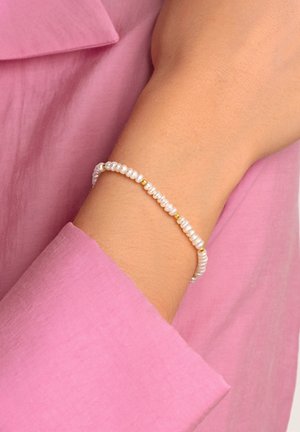 Bracciale di perle caratterizzato da piccole perle bianche intervallate da perline dorate, indossato su un polso, su uno sfondo di tessuto rosa.