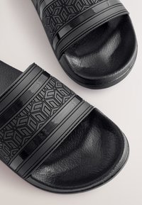 Sandali neri con fascia testurizzata a motivi geometrici, suola liscia e finitura opaca. Il design è minimalista e moderno.