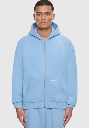 Hoodie zippé bleu clair en tissu doux, avec capuche, poches avant et poignets élastiques. Texture lisse avec une coupe décontractée.