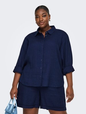 Femme aux cheveux tirés en arrière porte une chemise bleu marine à manches retroussées et un short assorti, tenant un sac à main bleu clair dans une main.