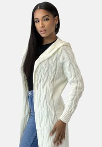 Cardigan lungo in maglia avorio con cappuccio, caratterizzato da motivi a treccia e sezioni a trama aperta. Il modello indossa un top nero e jeans blu.
