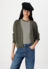 Olivovo zelený rebrovaný kardigán s piatimi gombíkmi, skombinovaný s tričkom v čierno-bielych prúžkoch a modrými džínsami s vysokým pásom. Čierna béretka na hlave.