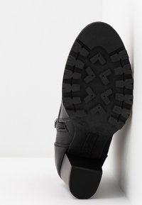 Bottine en cuir noir avec semelle à crampons robustes, talon bloc et fermeture éclair latérale, positionnée semelle vers le haut contre des murs blancs.