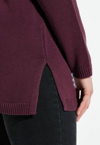 Suéter de punto burdeos con puños y dobladillo acanalados; presenta aberturas laterales, tejido texturizado y forro suave en el dobladillo. Llevado sobre pantalones negros.