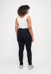 Jeans negros ajustados con bolsillos traseros, combinados con una blusa sin mangas blanca. El modelo está de espaldas, usando zapatillas grises. Textura de tela suave.