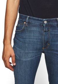 Jeans in denim blu scuro con una vestibilità slim. Le caratteristiche includono un bottone metallico, rivetti in ottone e una tasca frontale con dettagli di cucitura.