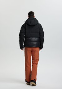 Chaqueta acolchada negra con capucha, que presenta un panel inferior brillante de color contraste. Combinada con pantalones impermeables naranjas y botas negras.