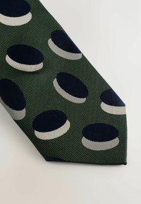 Groene stropdas van een gestructureerde stof met afwisselend donkere cirkels en witte accenten, gerangschikt in een patroon. De punt is spitz.