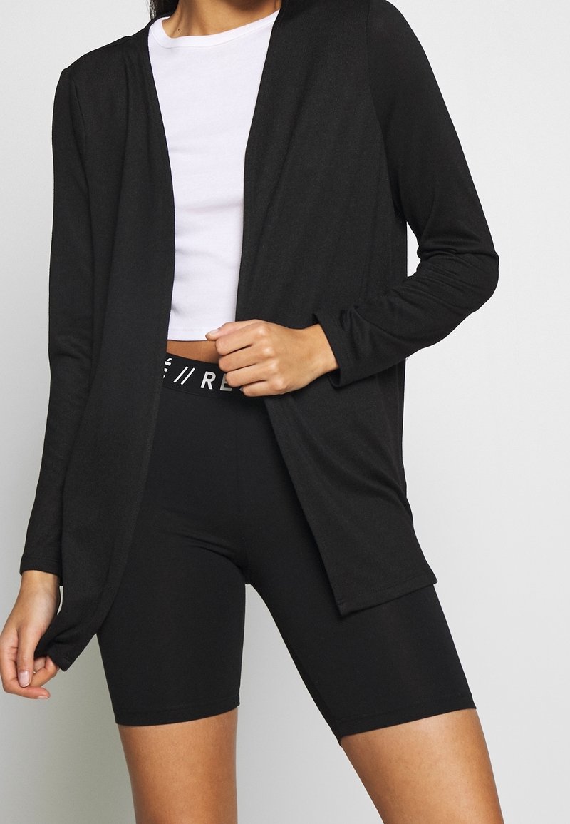 Černý cardigan s dlouhým rukávem, lehký materiál, otevřený design. Slučuje se s bílým crop topem a černými legínami s vysokým pasem a logem na pasu.