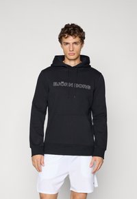 Jeune homme portant un hoodie noir avec le texte "Björn Borg" et un short blanc, debout contre un fond gris clair uni.
