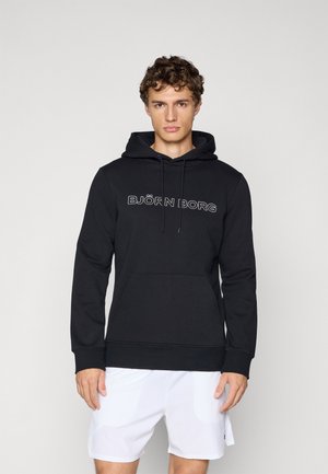 Ung mand iført en sort hoodie med "Björn Borg" tekst og hvide shorts, stående foran en ensfarvet lysegrå baggrund.