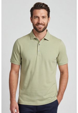 Man met kort bruin haar en baard, die een lichtgroen poloshirt met korte mouwen draagt, glimlachend met één hand in zijn zak.